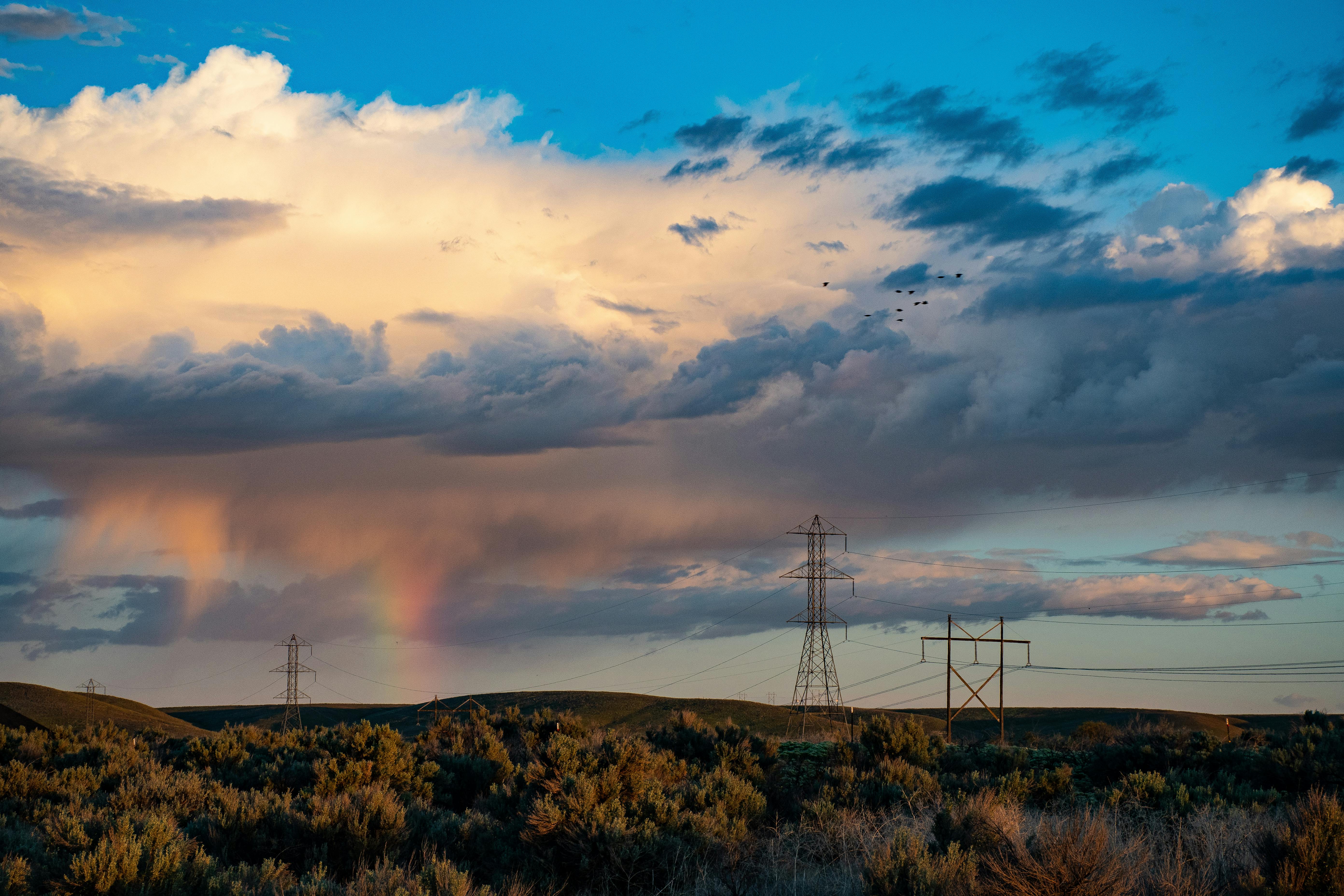 Dramatic sky with rainbow and powerlines over a natural landscape.